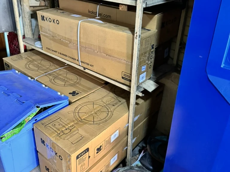 Boxed KOKO cookers and fuel canisters stacked on shelves in an agent shop's storeroom.